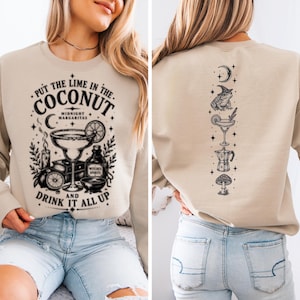 May include: A beige crewneck sweatshirt featuring a black graphic print. The front displays a margarita glass, lime, candle, and the text "PUT THE LIME IN THE COCONUT MIDNIGHT MARGARITAS AND DRINK IT ALL UP". The back shows a vertical design of a toad, cocktail, and mushroom.