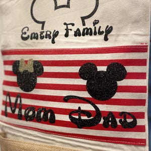 May include: A white canvas bag with red and white stripes. The bag has a Mickey Mouse head design with the words "Emery Family" at the top, "Mom" and "Dad" in the middle, and three Mickey Mouse heads at the bottom.