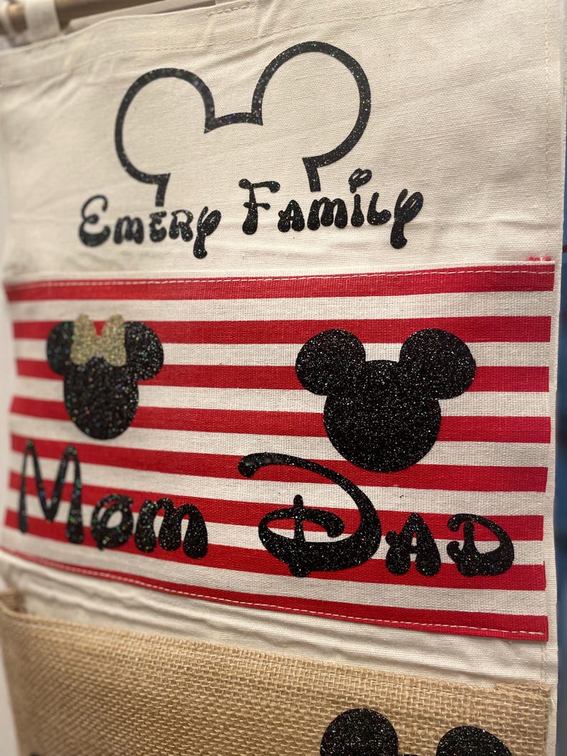 May include: A white canvas bag with red and white stripes. The bag has a Mickey Mouse head design with the words "Emery Family" at the top, "Mom" and "Dad" in the middle, and three Mickey Mouse heads at the bottom.