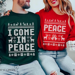 Puede incluir: Suéteres navideños a juego en verde bosque y rojo, con texto blanco y diseños festivos. El suéter verde dice "I COME IN PEACE" y el rojo "PEACE". Ambos incluyen renos, árboles y motivos de cajas de regalo.