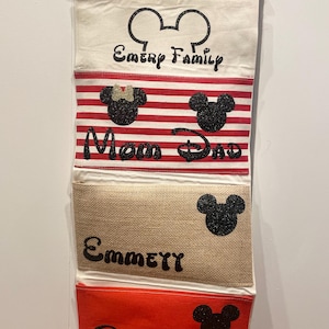 May include: A white canvas wall organizer with four pockets. The top pocket has a Mickey Mouse ear design and the text "Emery Family". The second pocket has a red and white striped design with a Mickey Mouse head and the text "Mom Dad". The third pocket is a beige burlap fabric with a Mickey Mouse head and the text "Emmett". The bottom pocket is red with a Mickey Mouse head and the text "Eli".