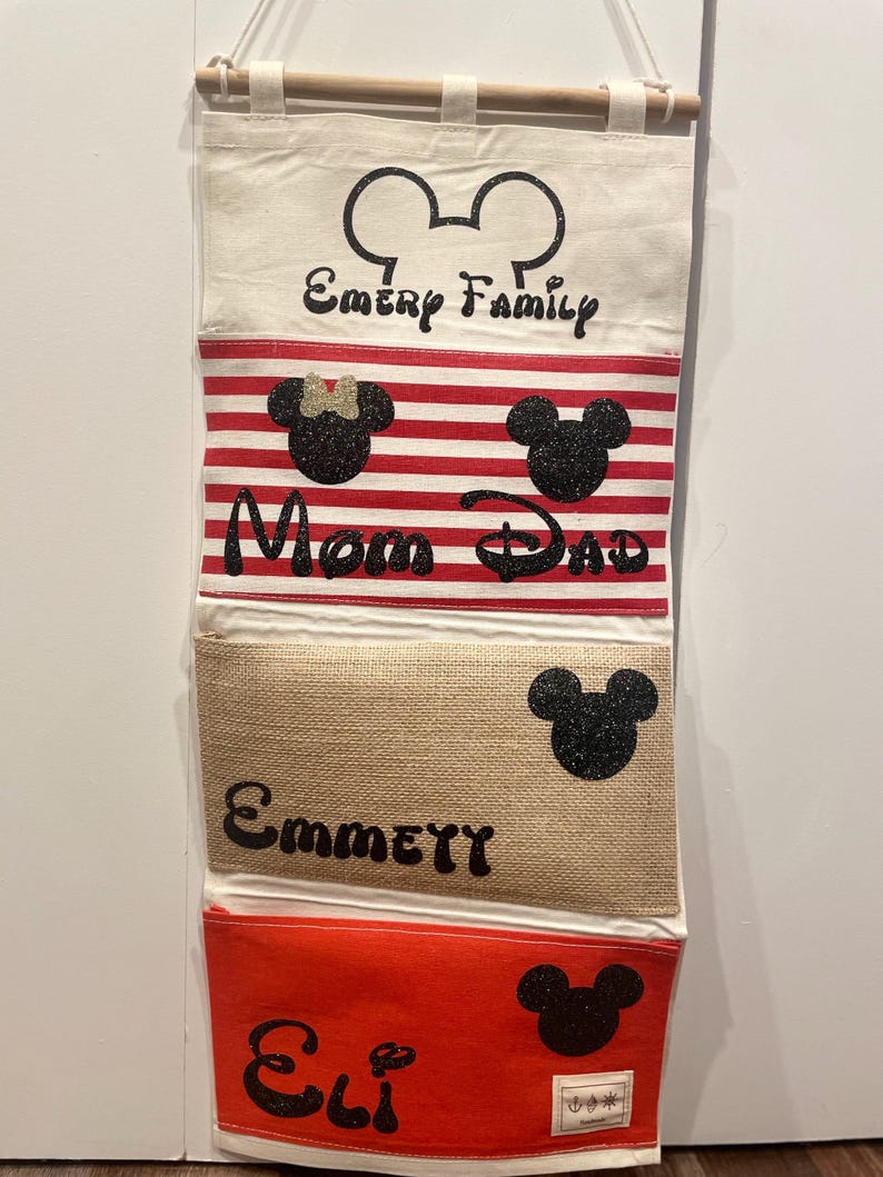 May include: A white canvas wall organizer with four pockets. The top pocket has a Mickey Mouse ear design and the text "Emery Family". The second pocket has a red and white striped design with a Mickey Mouse head and the text "Mom Dad". The third pocket is a beige burlap fabric with a Mickey Mouse head and the text "Emmett". The bottom pocket is red with a Mickey Mouse head and the text "Eli".