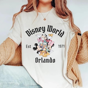 May include: White t-shirt with a black graphic of Mickey Mouse, Minnie Mouse, Donald Duck, Goofy, and Pluto. The graphic includes the text "Disney World Est 1971 Orlando".