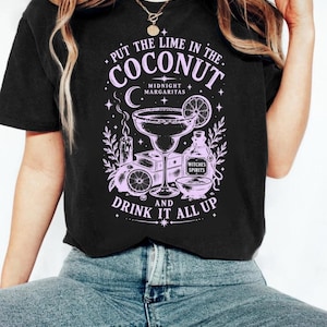 May include: A black t-shirt featuring a light purple graphic print of a margarita glass, a bottle labeled Witches Spirits, a candle, and citrus slices. The text reads Put the lime in the coconut, Midnight Margaritas, and Drink it all up.