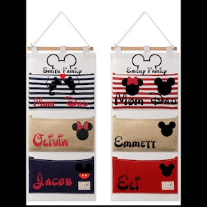 Puede incluir: Dos organizadores de pared de lona blanca con tres bolsillos cada uno. Los organizadores presentan un diseño de cabeza de Mickey Mouse y están personalizados con los nombres "Smith Family", "Emery Family", "Mom", "Dad", "Olivia", "Emmett", "Jacob" y "Eli".