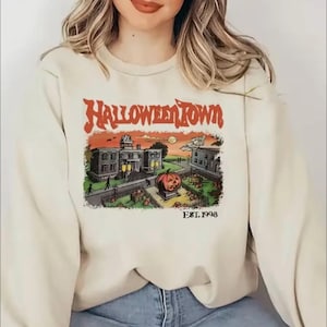 May include: A cream coloured sweatshirt with a graphic print of a cartoon town with a pumpkin in front of a building. The text "HALLOWEEN TOWN EST. 1998" is printed on the sweatshirt.