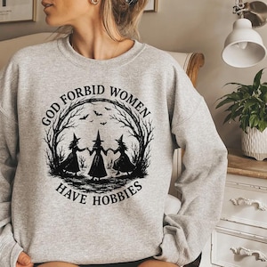 May include: A pale grey sweatshirt featuring a black graphic of three witches in a circle of trees, with bats flying above. The text reads "GOD FORBID WOMEN HAVE HOBBIES" in black.