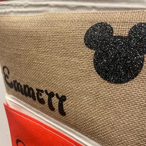 May include: A burlap storage bin with a black glitter Mickey Mouse head and the word "Emily" written in black. The bin is part of a set of bins with red and white striped fabric at the top.