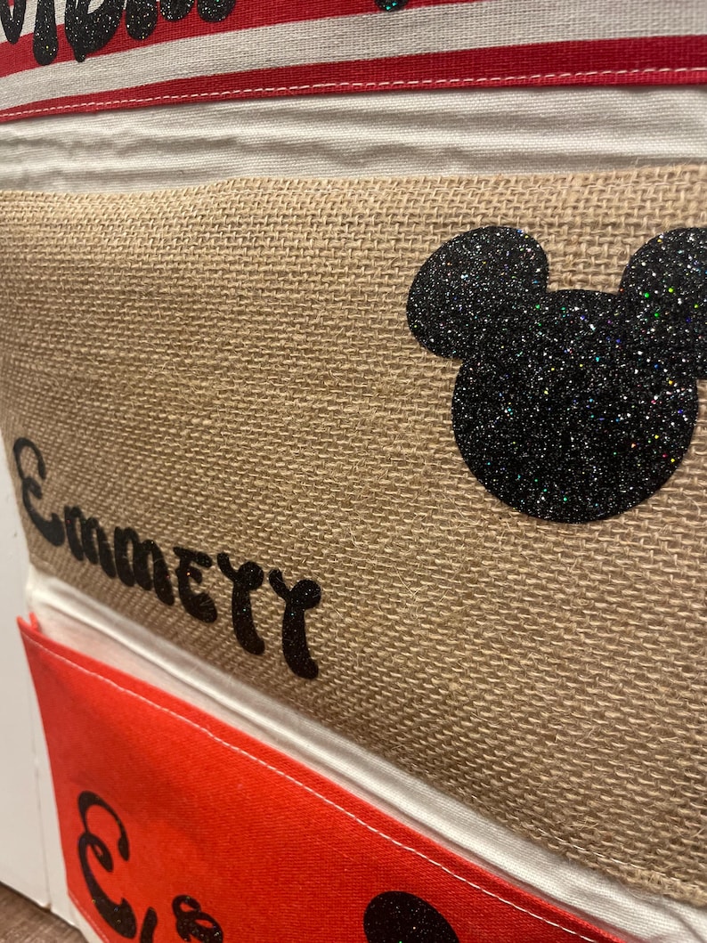 May include: A burlap storage bin with a black glitter Mickey Mouse head and the word "Emily" written in black. The bin is part of a set of bins with red and white striped fabric at the top.