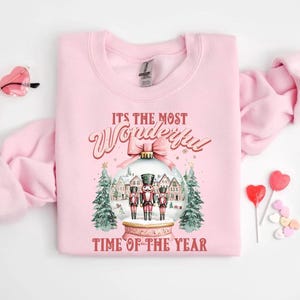May include: A pale pink sweatshirt with the phrase "It's the Most Wonderful Time of the Year." The design includes a snow globe with nutcrackers, trees, and houses. Heart-shaped sunglasses and sweets are also visible.