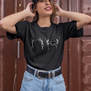 May include: A black t-shirt with a graphic of four skeletons dancing. The skeletons are white and are in different poses. The person wearing the shirt is also wearing a black cap and blue denim shorts.