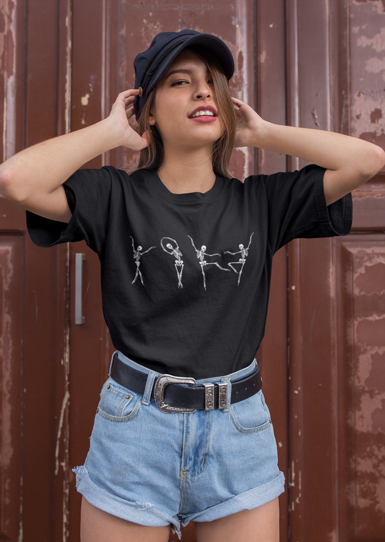 May include: A black t-shirt with a graphic of four skeletons dancing. The skeletons are white and are in different poses. The person wearing the shirt is also wearing a black cap and blue denim shorts.