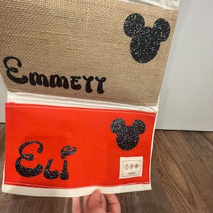 May include: A red and beige fabric wall organizer with two pockets. The top pocket has the name "EmmeTT" and a black glitter Mickey Mouse head. The bottom pocket has the name "Eli" and a black glitter Mickey Mouse head. The organizer has a white border and a red and white striped top.