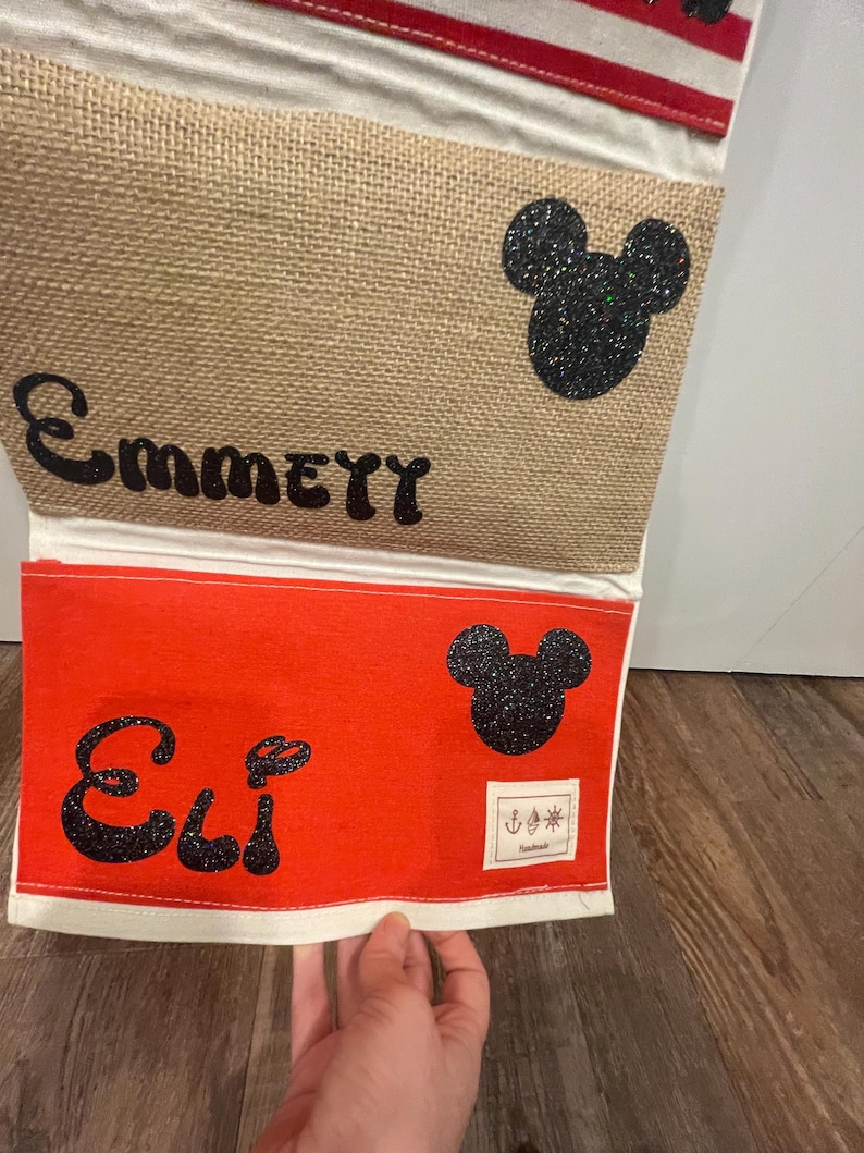 May include: A red and beige fabric wall organizer with two pockets. The top pocket has the name "EmmeTT" and a black glitter Mickey Mouse head. The bottom pocket has the name "Eli" and a black glitter Mickey Mouse head. The organizer has a white border and a red and white striped top.