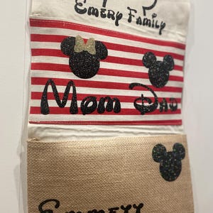 May include: A white canvas wall organizer with red and white striped fabric pockets. The top pocket has the text "Emery Family" and a black Mickey Mouse head. The second pocket has the text "Mom Dad" and a black Mickey Mouse head and a black Minnie Mouse head. The third pocket has the text "Emmey" and a black Mickey Mouse head.