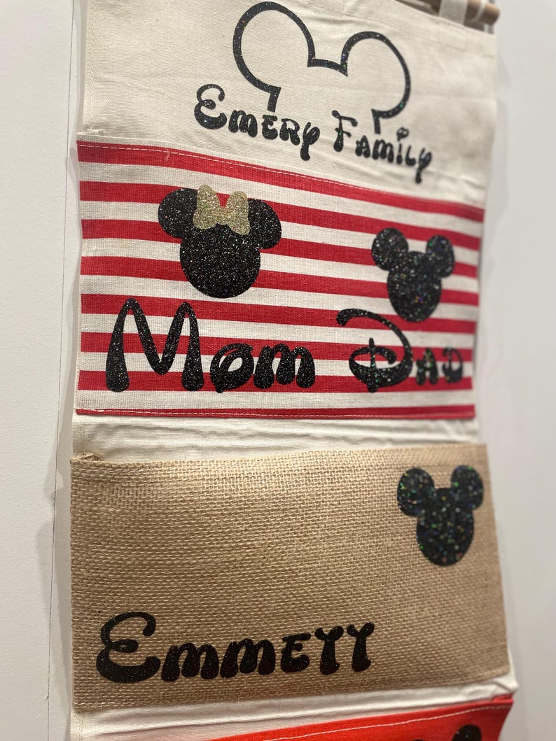 May include: A white canvas wall organizer with red and white striped fabric pockets. The top pocket has the text "Emery Family" and a black Mickey Mouse head. The second pocket has the text "Mom Dad" and a black Mickey Mouse head and a black Minnie Mouse head. The third pocket has the text "Emmey" and a black Mickey Mouse head.