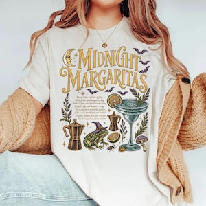 以下が含まれることがあります： ゴールドの文字で「Midnight Margaritas」と書かれたクリーム色のTシャツ。グラフィックには、三日月、コウモリ、マルガリータグラス、紫色の魔女の帽子をかぶったカエル、コーヒーポット、そしてポーションのレシピが書かれた小さなテキストが含まれています。