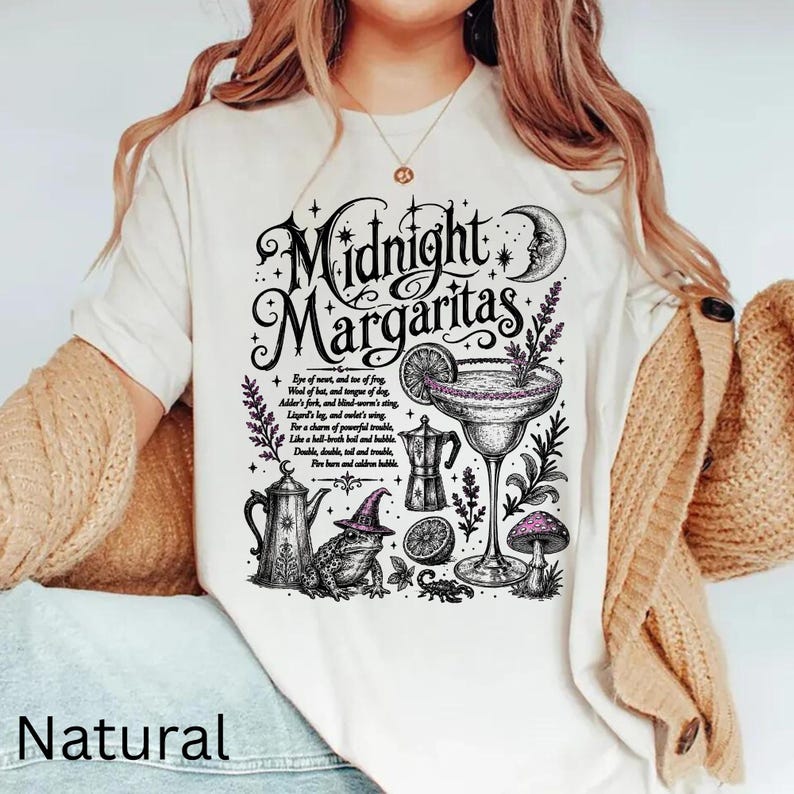 May include: A cream-colored t-shirt featuring a black graphic print with the text Midnight Margaritas, a crescent moon, a margarita glass, a frog wearing a witch hat, a coffee pot, a mushroom, and a scorpion, surrounded by stars and sprigs.