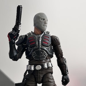 May include: Action figure of a masked character in a black and gray tactical suit, holding a black pistol. The figure has a gray mask, a detailed chest plate with red accents, and a utility belt. The figure is a collectible item.