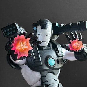 May include: War Machine action figure in a dynamic pose, featuring a black and silver armored suit. The figure has a white faceplate and is holding two energy blasts with pink and orange hues. The figure has a cannon on its back.
