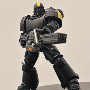 May include: A miniature figurine of a soldier in matte black armor with yellow accents on the helmet and shoulder. The figure is standing in a wide stance, holding a large, multi-barreled gray weapon with both hands, pointed forward.