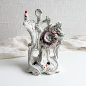 May include: A white ceramic decorative vase featuring a bird and nest design. The vase has a tree-like structure with a bird perched on top and a nest with eggs in the center. Accents of gold and red add detail to the piece.