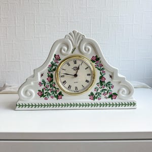 May include: White ceramic mantel clock with a gold-coloured clock face and Roman numerals. The clock is decorated with pink floral and green leaf patterns. The clock face reads "Quartz".