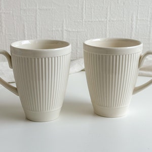May include: Two cream-coloured ceramic mugs with vertical ribbed detailing. Each mug has a curved handle and a slightly flared shape. The mugs are set against a white background, suggesting a clean and minimalist aesthetic.