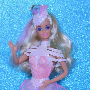 May include: A doll with long blonde hair featuring pastel highlights, wearing a pink bodice with gold polka dots, a white iridescent scalloped collar, and a matching headpiece. The doll has blue eyes, pink lips, and a sheer purple skirt overlay.