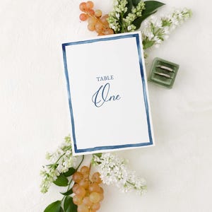 May include: A white card with a blue border and the text "TABLE One" in blue calligraphy. The card is surrounded by white flowers and green leaves.