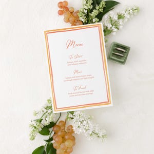 Op de afbeelding: Een aquarelmenukaart met een perzik- en oranje rand. Het menu is geschreven in cursief schrift en vermeldt de gangen: Om te beginnen, Hoofdgerecht en Om te eindigen. De menu-items zijn: Tomaat, basilicum, komkommer met balsamico-reductie; Zeebaars met citroenbotersaus, zuurdesembrood en verse rucola; Verse seizoensgebonden bessen met slagroom en chocoladeschilfers.