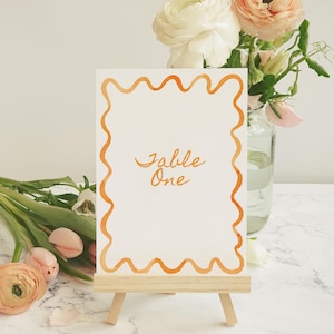 Könnte beinhalten: Eine weiße Karte mit einem orangefarbenen, wellenförmigen Rand und dem Text "Table One" in orangefarbener Kursivschrift. Die Karte steht auf einem kleinen Holzständer.