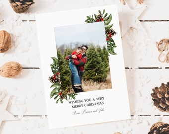 Peut inclure: Une carte de Noël avec une photo d'un père et de son fils dans une plantation de sapins de Noël. La carte a une bordure florale verte et rouge avec des baies et des pommes de pin. Le texte sur la carte dit "Wishing you a very Merry Christmas from Darren and Jake".