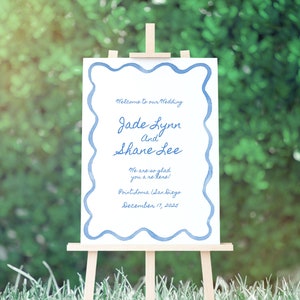 May include: A white wedding sign with blue watercolor border, announcing the wedding of Jade Lynn and Shane Lee. The sign also includes the date and location of the wedding: December 17, 2025, Point Loma, San Diego.