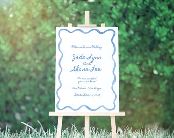 Op de afbeelding: Een wit huwelijksbord met een blauwe aquarelrand, dat de bruiloft van Jade Lynn en Shane Lee aankondigt. Het bord bevat ook de datum en locatie van de bruiloft: 17 december 2025, Point Loma, San Diego.
