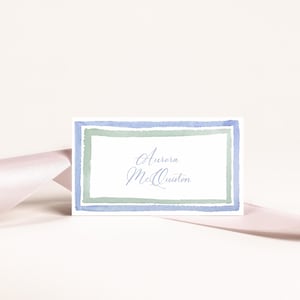Puede incluir: Una tarjeta de lugar blanca con un borde de acuarela azul y verde. El nombre "Aurora McQuiston" está escrito en cursiva dentro del borde.