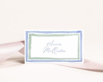 May include: A white place card with a blue and green watercolour border. The name "Aurora McQuiston" is written in cursive inside the border.