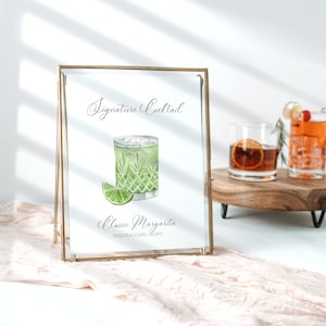 May include: A gold-framed sign with the text "Signature Cocktail" and a watercolor illustration of a classic margarita. The text below the illustration reads "Classic Margarita Tequila | Lime | Agave".