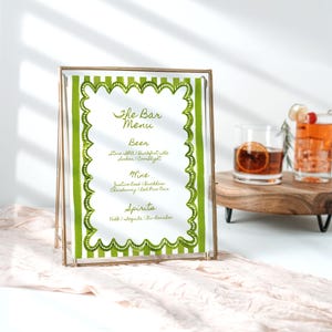 Puede incluir: Un menú de bar enmarcado en un marco dorado. El menú presenta detalles a rayas verdes y blancas y enumera cerveza, vino y licores. Dos cócteles se ven en una bandeja de madera en el fondo.