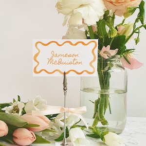 Puede incluir: Una tarjeta blanca con un borde naranja y el texto "Jameson McQuiston" en escritura naranja. La tarjeta está unida a un clip plateado y una cinta blanca. La tarjeta está delante de un jarrón de flores blancas y rosas.