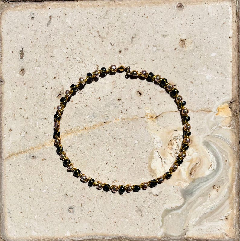 K&ouml;nnte beinhalten: Ein kreisf&ouml;rmiges Armband aus einem d&uuml;nnen goldfarbenen Draht, das mit einem abwechselnden Muster aus kleinen, glatten schwarzen Perlen und metallisch silberfarbenen Perlen besetzt ist, die durchgehend auf dem Draht aufgereiht sind.