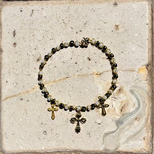 K&ouml;nnte beinhalten: Ein kreisf&ouml;rmiges Perlenarmband mit abwechselnd schwarzen und durchscheinend wei&szlig;en Perlen auf einem goldfarbenen Draht. Drei goldfarbene Metallkreuz-Anh&auml;nger h&auml;ngen an der Unterseite, mit einem gr&ouml;&szlig;eren, verzierten Kreuz in der Mitte.
