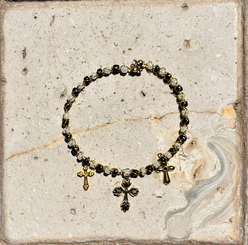 K&ouml;nnte beinhalten: Ein kreisf&ouml;rmiges Perlenarmband mit abwechselnd schwarzen und durchscheinend wei&szlig;en Perlen auf einem goldfarbenen Draht. Drei goldfarbene Metallkreuz-Anh&auml;nger h&auml;ngen an der Unterseite, mit einem gr&ouml;&szlig;eren, verzierten Kreuz in der Mitte.