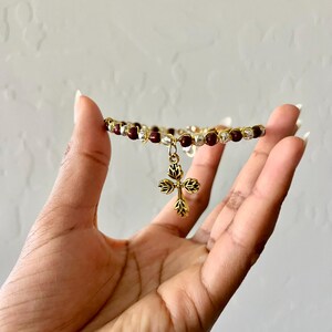 K&ouml;nnte beinhalten: Ein Perlenarmband mit abwechselnd dunkelroten und silberfarbenen Perlen auf einem goldfarbenen Draht. Ein goldfarbener Metallanh&auml;nger in Kreuzform mit einem stilisierten Blatt- oder Bl&uuml;tenblattmuster h&auml;ngt mittig am Armband.