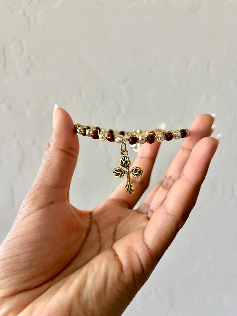K&ouml;nnte beinhalten: Ein Perlenarmband mit abwechselnd dunkelroten und silberfarbenen Perlen auf einem goldfarbenen Draht. Ein goldfarbener Metallanh&auml;nger in Kreuzform mit einem stilisierten Blatt- oder Bl&uuml;tenblattmuster h&auml;ngt mittig am Armband.