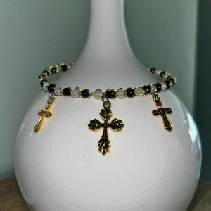 K&ouml;nnte beinhalten: Ein Perlenarmband mit abwechselnd schwarzen und wei&szlig;en Perlen auf einem d&uuml;nnen Draht. Das Armband ist mit drei goldfarbenen Metallkreuz-Anh&auml;ngern verziert, wobei sich ein gr&ouml;&szlig;eres, verziertes Kreuz in der Mitte und zwei kleinere Kreuze befinden.