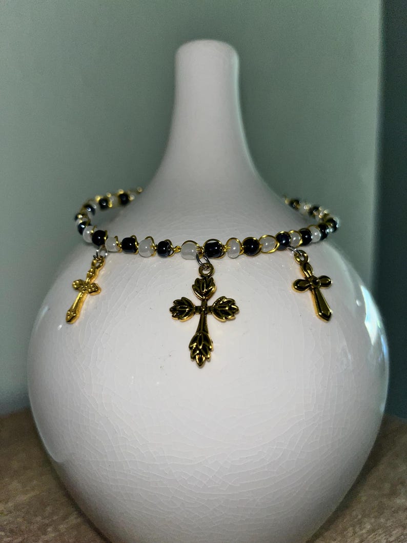 K&ouml;nnte beinhalten: Ein Perlenarmband mit abwechselnd schwarzen und wei&szlig;en Perlen auf einem d&uuml;nnen Draht. Das Armband ist mit drei goldfarbenen Metallkreuz-Anh&auml;ngern verziert, wobei sich ein gr&ouml;&szlig;eres, verziertes Kreuz in der Mitte und zwei kleinere Kreuze befinden.