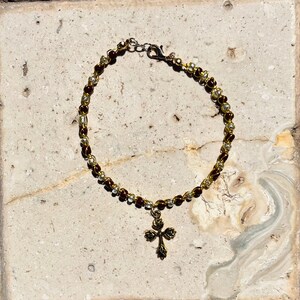 K&ouml;nnte beinhalten: Ein Perlenarmband mit abwechselnd dunkelbraunen und goldfarbenen Metallperlen auf einem Draht. Das Armband verf&uuml;gt &uuml;ber einen goldfarbenen Metallanh&auml;nger in Kreuzform mit einem dekorativen Blattmuster und einen Karabinerverschluss.