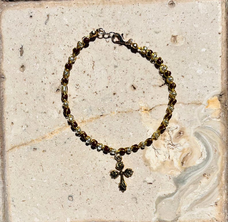 K&ouml;nnte beinhalten: Ein Perlenarmband mit abwechselnd dunkelbraunen und goldfarbenen Metallperlen auf einem Draht. Das Armband verf&uuml;gt &uuml;ber einen goldfarbenen Metallanh&auml;nger in Kreuzform mit einem dekorativen Blattmuster und einen Karabinerverschluss.