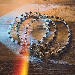 K&ouml;nnte beinhalten: Drei zarte Armb&auml;nder mit kleinen, abwechselnd dunklen und hellen Perlen auf d&uuml;nnem Metalldraht. Jedes Armband hat einen kleinen Metallanh&auml;nger, wobei eines ein Kreuz und ein anderes ein blumen- oder blattartiges Design zeigt.
